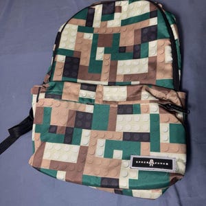 May include: A backpack with a pixelated pattern in shades of green, brown, tan, and black. The backpack has a black zipper and straps. A small rectangular tag on the front reads "SPACE JUNKE" with a graphic.