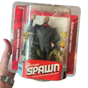 Peut inclure: Figurine d'action emballée de la série "The Adventures of Spawn". La figurine est dans un étui en plastique transparent, avec un dos rouge et blanc. La figurine représente un homme chauve en costume, avec deux monstres verts et d'autres accessoires.