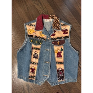 May include: A blue denim vest featuring a button-up front and a collar with contrasting red and brown patterned fabric. The front panels are decorated with vertical strips of beige fabric displaying illustrations of angels, stars, and moons.