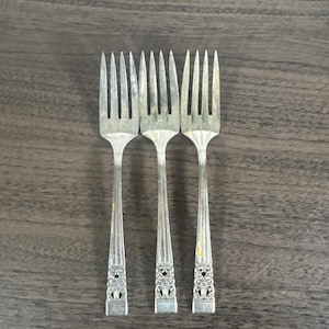 May include: Three silver forks with decorative floral patterns on the handles are displayed on a dark wooden surface. The forks have multiple tines and an ornate design near the handle's base.