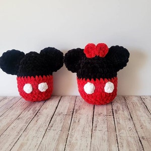 May include: Two plush Mickey Mouse and Minnie Mouse inspired figures. The figures have black ears, red bodies with white circles, and a red bow on one. The figures are handmade and sit on a light wood surface.