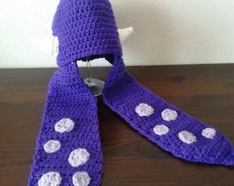 Könnte beinhalten: Eine violette Häkelmütze mit zwei langen, herabhängenden Seitenteilen und zwei kleinen, weißen, spitzen Ohren an den Seiten. Die langen Seitenteile sind mit mehreren hellvioletten, kreisförmigen Flecken verziert, die an Tentakel aus Garn erinnern.