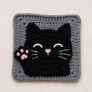May include: A square crochet coaster featuring a black cat face with a raised paw. The cat has white embroidered eyes, a pink nose, and grey whiskers. The paw pad is pink. The cat is centered on a textured grey square background with a border.
