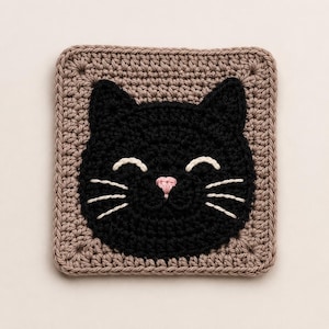 May include: A square crochet coaster featuring a black cat face centered on a light brown background. The cat has embroidered white curved eyes, white whiskers, and a small pink triangular nose, all worked in a detailed yarn stitch pattern.