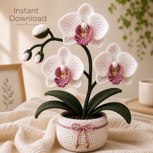 Pode incluir: Uma orquídea de crochê com pétalas brancas, um centro rosa e amarelo e folhas verde-escuras em um vaso de crochê branco com um laço rosa. O texto Instant Download está visível no canto superior esquerdo deste arranjo floral têxtil.