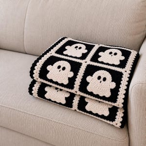 Peut inclure: Une couverture en crochet pliée présentant un motif répétitif de fantômes couleur crème centrés dans des carrés noirs. La couverture est bordée d'une bordure festonnée couleur crème, chaque fantôme ayant deux yeux circulaires sombres.