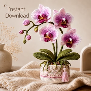Pode incluir: Uma orquídea de crochê com pétalas rosa e brancas, folhas verde-escuras e vários botões pequenos em uma haste. A planta está em um vaso de crochê texturizado na cor creme, adornado com uma pequena borla rosa. Texto: Instant Download.