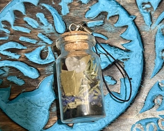 May include: A miniature glass bottle with a cork stopper, containing dried herbs, crystals, and other natural elements. It hangs from a silver clasp and black cord. The background is a wooden surface with a blue carved design.