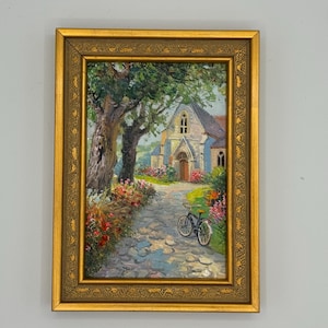 May include: A framed oil painting depicts a church with a cobblestone path and a bicycle. The artwork features a gold frame and a vibrant color palette of greens, blues, and reds. The scene is set in a picturesque landscape.