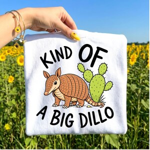 May include: White t-shirt with a cartoon armadillo and cactus graphic. The text reads "KIND OF A BIG DILLO" in black. The armadillo is brown and the cactus is green. The background is a field of sunflowers.