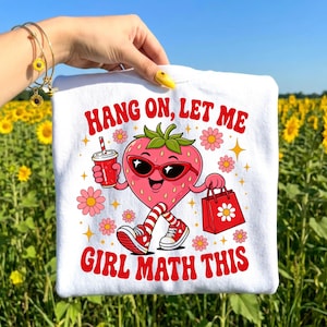 May include: White t-shirt with a cartoon strawberry wearing sunglasses, holding a drink and shopping bag. The text "HANG ON, LET ME GIRL MATH THIS" is printed in red. Pink flowers and stars surround the strawberry design.