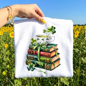 May include: White t-shirt featuring a stack of books with a teacup and shamrocks. The books are green and red with gold accents, and the teacup has shamrock designs. A green ribbon is tied around the books.
