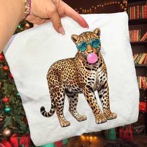 May include: White pillow with a leopard print design. The leopard is wearing heart-shaped sunglasses and blowing a bubblegum bubble. The leopard has a brown and black spotted pattern.