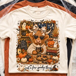 May include: A cream-coloured t-shirt featuring a graphic of a Highland cow wearing a bow and heart-shaped sunglasses, holding a pumpkin mug. The design includes pumpkins, ghosts, books labelled Pumpkin Spells, Ghost Stories, Witches Brew, and the text "STAY COZY AND SPOOKY" and "just a few spooky things".