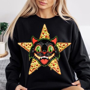 May include: Black sweatshirt featuring a cartoon cat face with green eyes and a red mouth, surrounded by a pizza slice star design. The cat has a mischievous grin. The sweatshirt is a casual, comfortable style.