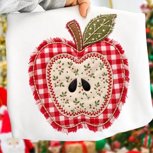 May include: A white sweatshirt featuring a large, centered apple applique. The apple has a red and white gingham border with frayed edges, a cream-colored center with a small floral pattern, two black seeds, and a green leaf with white dots.