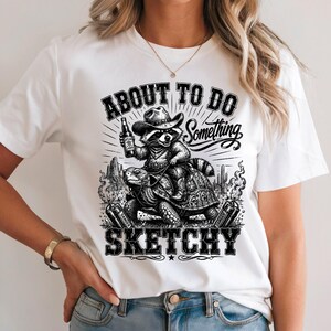 May include: White t-shirt featuring a black and white illustration of a raccoon in a cowboy hat, riding a turtle and holding a beer bottle. The text reads "ABOUT TO DO SOMETHING SKETCHY".