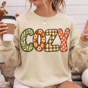 May include: A beige crewneck sweatshirt featuring the word COZY printed in large letters. Each letter has a distinct pattern: the C has green leaves, the O has white daisies on orange, the Z has a tan plaid, and the Y has white polka dots.