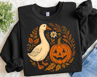 May include: Black sweatshirt featuring a cream-colored goose and a jack-o'-lantern. The goose and pumpkin are surrounded by fall foliage in shades of orange and brown. A white flower is also included in the design. The sweatshirt is a fall-themed item.