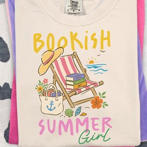 Op de afbeelding: Een crèmekleurig t-shirt met een afbeelding van een strandstoel, zonnehoed, draagtas met anker, boeken, bloemen en schelpen. De tekst luidt BOOKISH SUMMER Girl in geel, roze en groen. Het t-shirt is voorzien van een Comfort Colors label.
