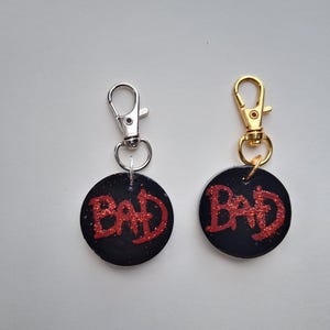 May include: Two keychains with the word "BAD" in red glitter on a black circular background. One keychain has a silver clasp, and the other has a gold clasp. The keychains are displayed against a white background.