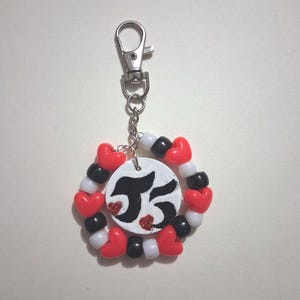 May include: A keychain featuring a circular white pendant with black stylized text and two small red glitter hearts. The pendant is surrounded by a ring of alternating red heart-shaped beads, white round beads, and black round beads on a metal chain.