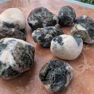 May include: A selection of smooth, natural stones. The stones vary in size and shape, with a mix of white, grey, and green coloration. Some stones have a mottled appearance, while others are more solid in colour. The stones are arranged on a reddish-brown surface.