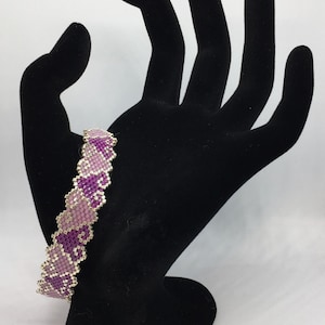 May include: A purple and pink beaded bracelet with a heart design. The bracelet is on a black hand-shaped display stand.