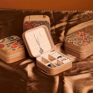 Puede incluir: Cuatro joyeros de corcho con diseños florales y estampados. Una caja está abierta, revelando compartimentos para anillos y un collar. Las cajas tienen un exterior de corcho natural y un interior beige, con cierre de cremallera.