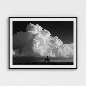 May include: Black and white framed print featuring a dramatic cloudscape. A person on a motorcycle is silhouetted against the horizon. The image captures a sense of vastness and movement, with the clouds dominating the sky.