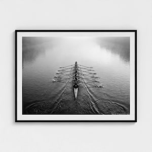 Może przedstawiać: Czarno-biała oprawiona grafika przedstawiająca drużynę wioślarską na zamglonym jeziorze. Łódź z ośmioma wioślarzami i sternikiem przecina wodę, pozostawiając za sobą ślad. Zdjęcie wykonane z góry, podkreślające symetrię i pracę zespołową.