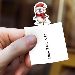 May include: A hand holds a white paper note with the text "Dein Text hier". A Christmas-themed penguin sticker with a red hat and scarf is attached to the note. The number "18" is printed on the sticker.