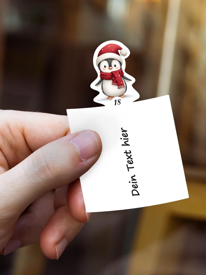 May include: A hand holds a white paper note with the text "Dein Text hier". A Christmas-themed penguin sticker with a red hat and scarf is attached to the note. The number "18" is printed on the sticker.