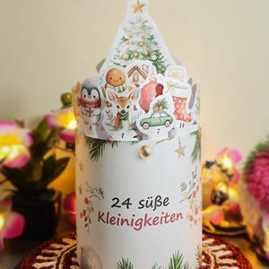 Puede incluir: Un calendario de adviento cil&iacute;ndrico con una parte superior decorativa en forma de &aacute;rbol. El calendario presenta ilustraciones festivas de un ping&uuml;ino, un ciervo, un hombre de jengibre y un coche. El texto "24 s&uuml;&szlig;e Kleinigkeiten" est&aacute; impreso en el lateral.