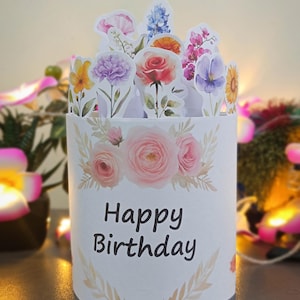以下が含まれることがあります： 白い円筒形の誕生日カード。花柄のデザインと黒字で「Happy Birthday」の文字が書かれています。カードは、バラ、カーネーション、ヒマワリなど、水彩画の花の切り抜きで飾られています。全体的なテーマは、お祝いと花です。