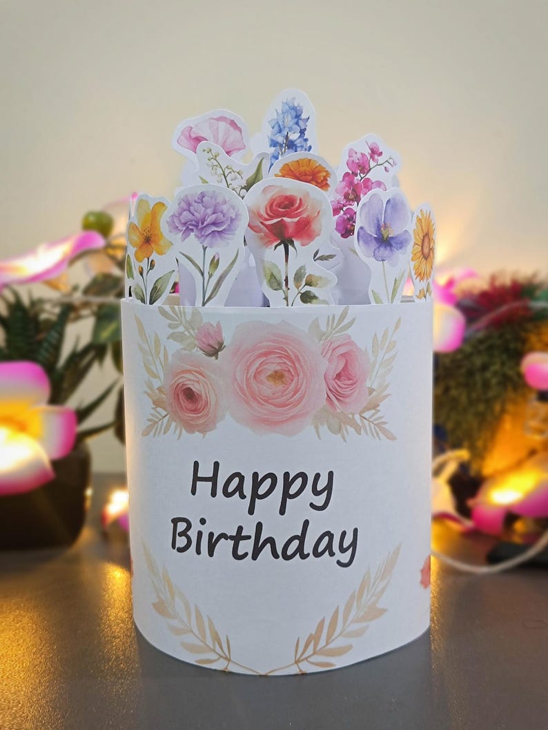 Puede incluir: Una tarjeta de cumplea&ntilde;os cil&iacute;ndrica blanca con un dise&ntilde;o floral y las palabras "Happy Birthday" en negro. La tarjeta est&aacute; adornada con recortes de flores de acuarela, incluyendo rosas, claveles y girasoles. El tema general es festivo y floral.