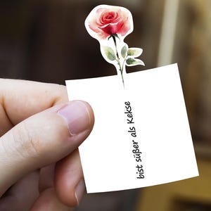 Puede incluir: Una pegatina de estilo acuarela con una rosa roja y rosa con hojas verdes. La rosa est&aacute; unida a un cuadrado blanco con el texto "bist s&uuml;&szlig;er als Kekse" en negro, sostenido por una mano.