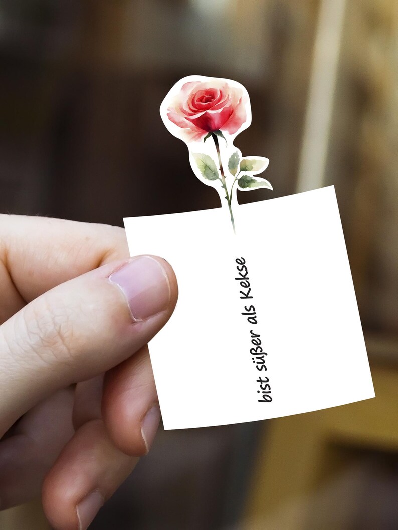 Puede incluir: Una pegatina de estilo acuarela con una rosa roja y rosa con hojas verdes. La rosa est&aacute; unida a un cuadrado blanco con el texto "bist s&uuml;&szlig;er als Kekse" en negro, sostenido por una mano.