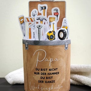 May include: A brown cylindrical container with a toolbox theme, featuring various tool cutouts like a hammer, saw, and tape measure. The container has the word "Papa" in script and German text.