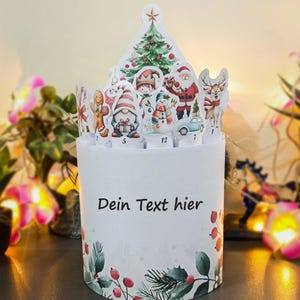 May include: A white Christmas advent calendar with a festive design. The top features a Christmas tree, Santa Claus, and other holiday characters. The base has the text "Dein Text hier" and is decorated with holly and berries.