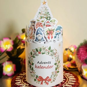 Puede incluir: Un calendario de Adviento cil&iacute;ndrico con un dise&ntilde;o festivo. El calendario presenta una corona con las palabras "Advents kalender" y un lazo rojo. Est&aacute; rematado con recortes con tem&aacute;tica navide&ntilde;a, que incluyen un &aacute;rbol de Navidad, mu&ntilde;ecos de nieve y un gnomo.