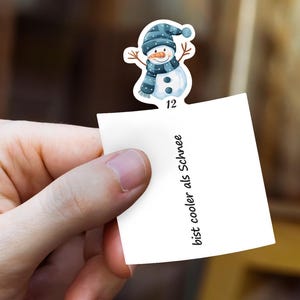 Puede incluir: Una mano sostiene un papel blanco con el texto "bist cooler als Schnee". Una pegatina de mu&ntilde;eco de nieve con gorro, bufanda y botones azules est&aacute; adherida al papel. El mu&ntilde;eco de nieve tiene una nariz de zanahoria y brazos de ramita. El n&uacute;mero 12 est&aacute; debajo.