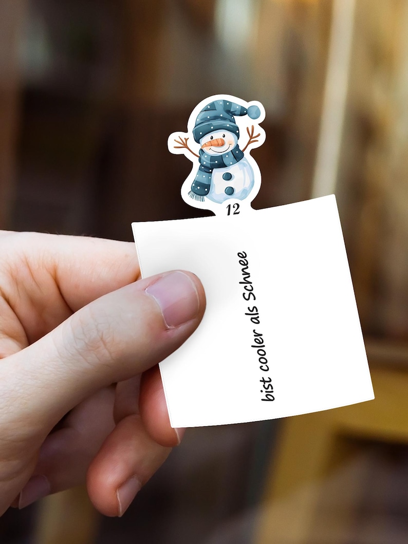 Puede incluir: Una mano sostiene un papel blanco con el texto "bist cooler als Schnee". Una pegatina de mu&ntilde;eco de nieve con gorro, bufanda y botones azules est&aacute; adherida al papel. El mu&ntilde;eco de nieve tiene una nariz de zanahoria y brazos de ramita. El n&uacute;mero 12 est&aacute; debajo.