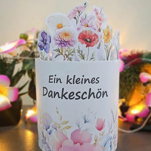 Puede incluir: Una caja de regalo cil&iacute;ndrica blanca con un dise&ntilde;o floral y el texto "Ein kleines Dankesch&ouml;n". La caja est&aacute; adornada con flores de estilo acuarela en rosa, morado y azul. La parte superior presenta formas de flores recortadas.