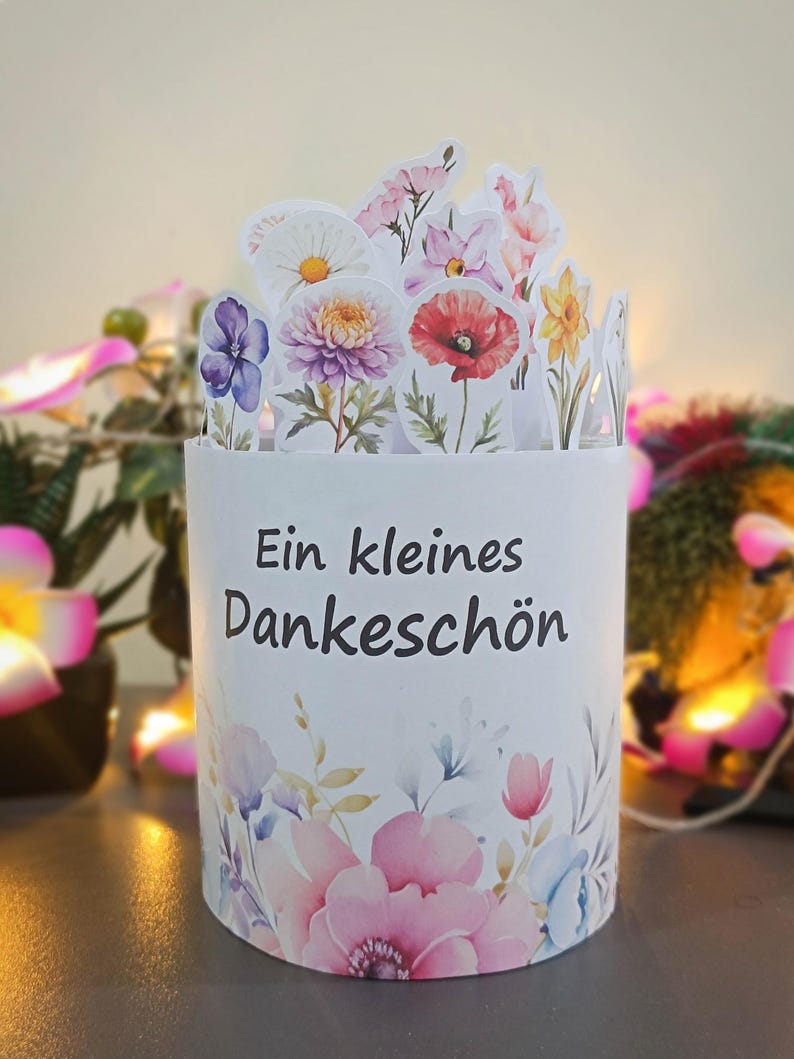 Puede incluir: Una caja de regalo cil&iacute;ndrica blanca con un dise&ntilde;o floral y el texto "Ein kleines Dankesch&ouml;n". La caja est&aacute; adornada con flores de estilo acuarela en rosa, morado y azul. La parte superior presenta formas de flores recortadas.