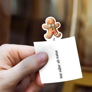 May include: A gingerbread man sticker with a smiling face, bow tie, and colorful accents is attached to a white note. The note has the number 2 and the text "bist s&uuml;&szlig;er als Kekse" in black.