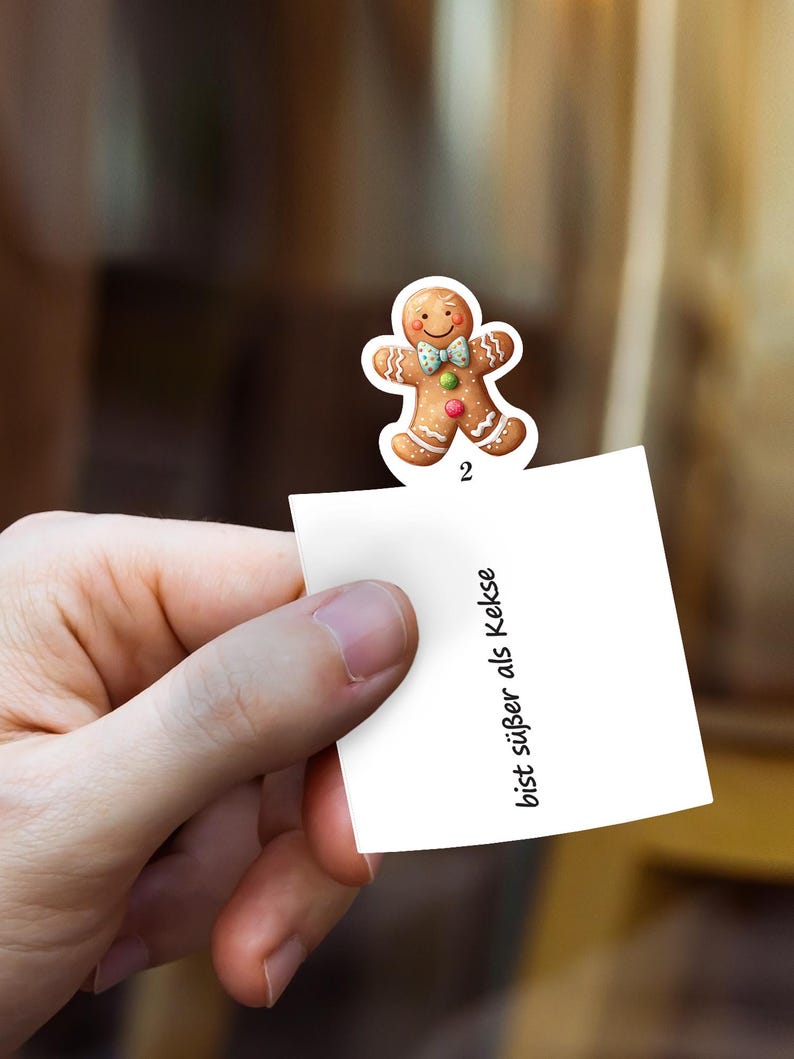 May include: A gingerbread man sticker with a smiling face, bow tie, and colorful accents is attached to a white note. The note has the number 2 and the text "bist s&uuml;&szlig;er als Kekse" in black.