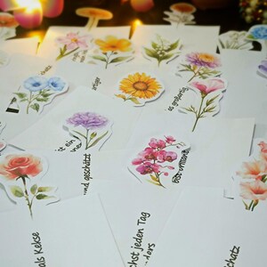 Puede incluir: Un conjunto de tarjetas blancas con pegatinas florales de acuarela, incluyendo rosas, girasoles y orqu&iacute;deas. Las tarjetas tienen texto en alem&aacute;n, como "bist s&uuml;&szlig;er als Kekse" y "bist ein Schatz". El fondo est&aacute; suavemente iluminado.