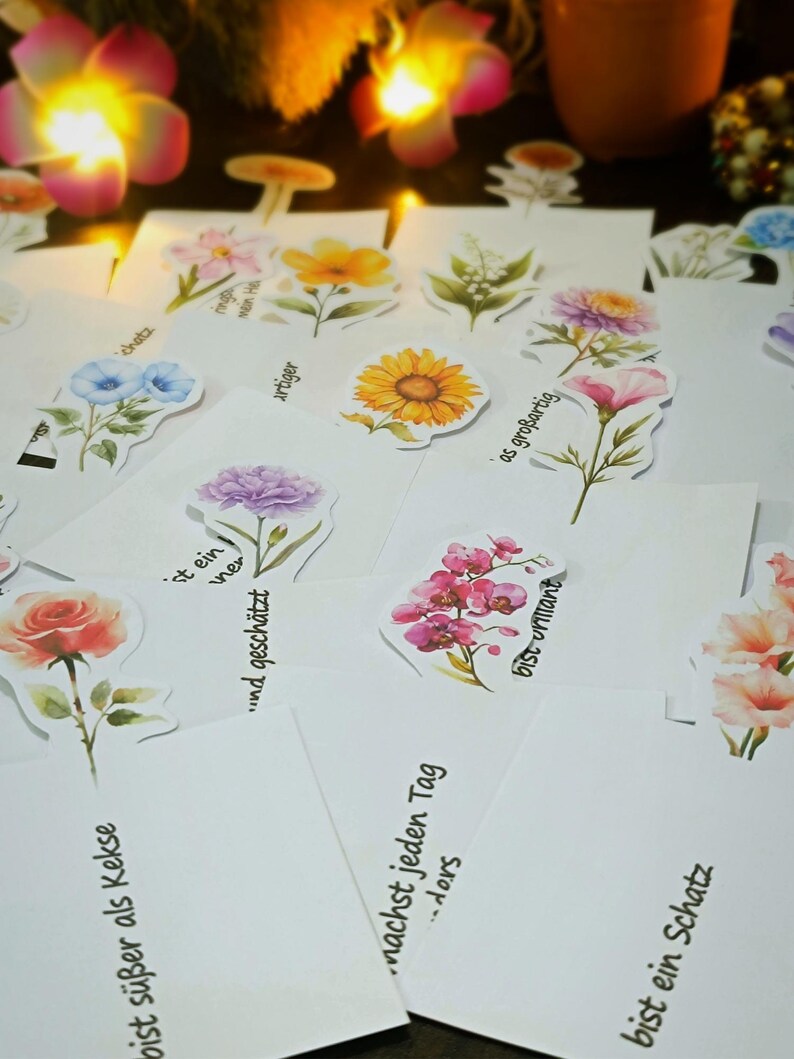Puede incluir: Un conjunto de tarjetas blancas con pegatinas florales de acuarela, incluyendo rosas, girasoles y orqu&iacute;deas. Las tarjetas tienen texto en alem&aacute;n, como "bist s&uuml;&szlig;er als Kekse" y "bist ein Schatz". El fondo est&aacute; suavemente iluminado.