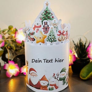 May include: A white Christmas advent calendar with a cylindrical base and a decorative top. The base features Santa Claus, snowmen, and polar bears. The top has cutouts of Christmas trees, stars, and other festive elements. The text "Dein Text hier" is printed on the base.